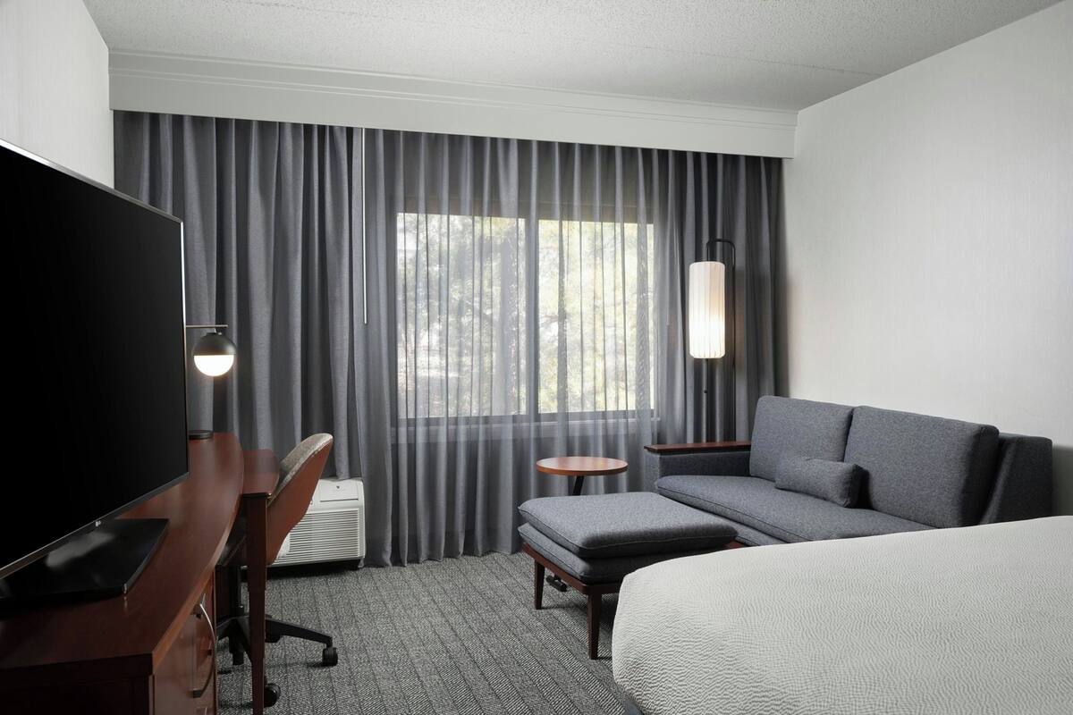 A hotel room with a bed, a sofa, a desk with a TV, a chair, a small table, and a window with curtains.