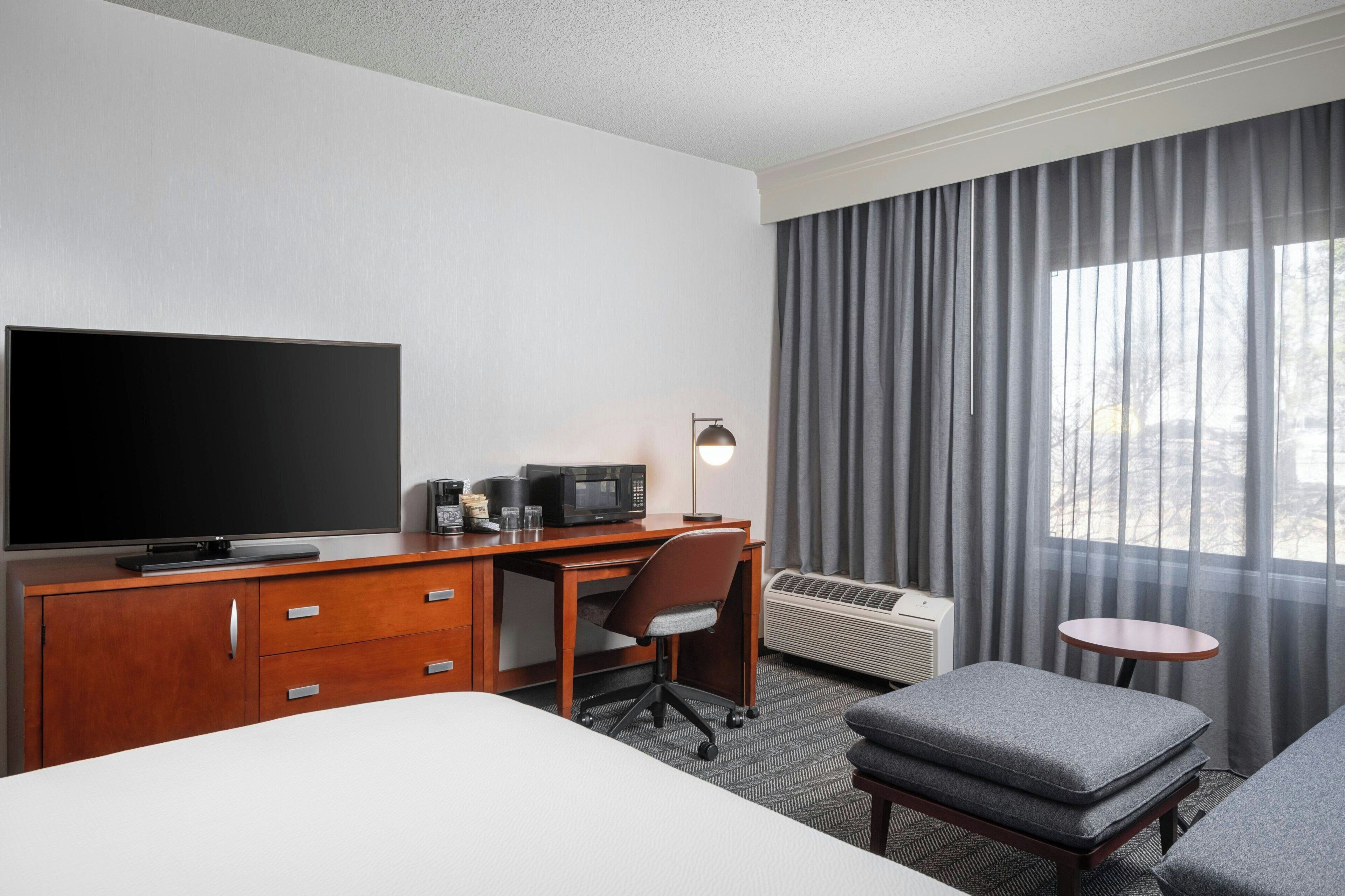 Ein Hotelzimmer mit einem Bett, einem Schreibtisch mit Flachbildfernseher, einem Sessel, einem kleinen runden Tisch und einem Fenster mit Vorhängen.