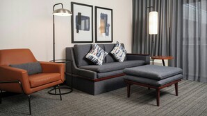 A modern hotel room with a sofa, armchair, ottoman, floor lamp, and abstract art on the wall.