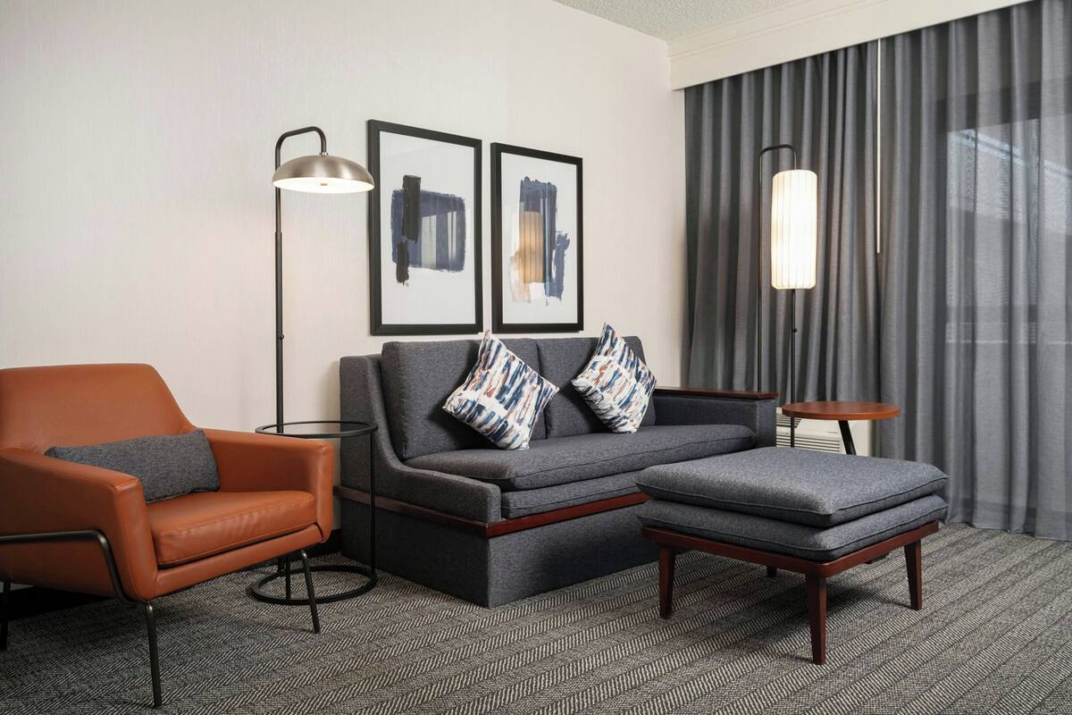 A modern hotel room with a sofa, armchair, ottoman, floor lamp, and abstract art on the wall.