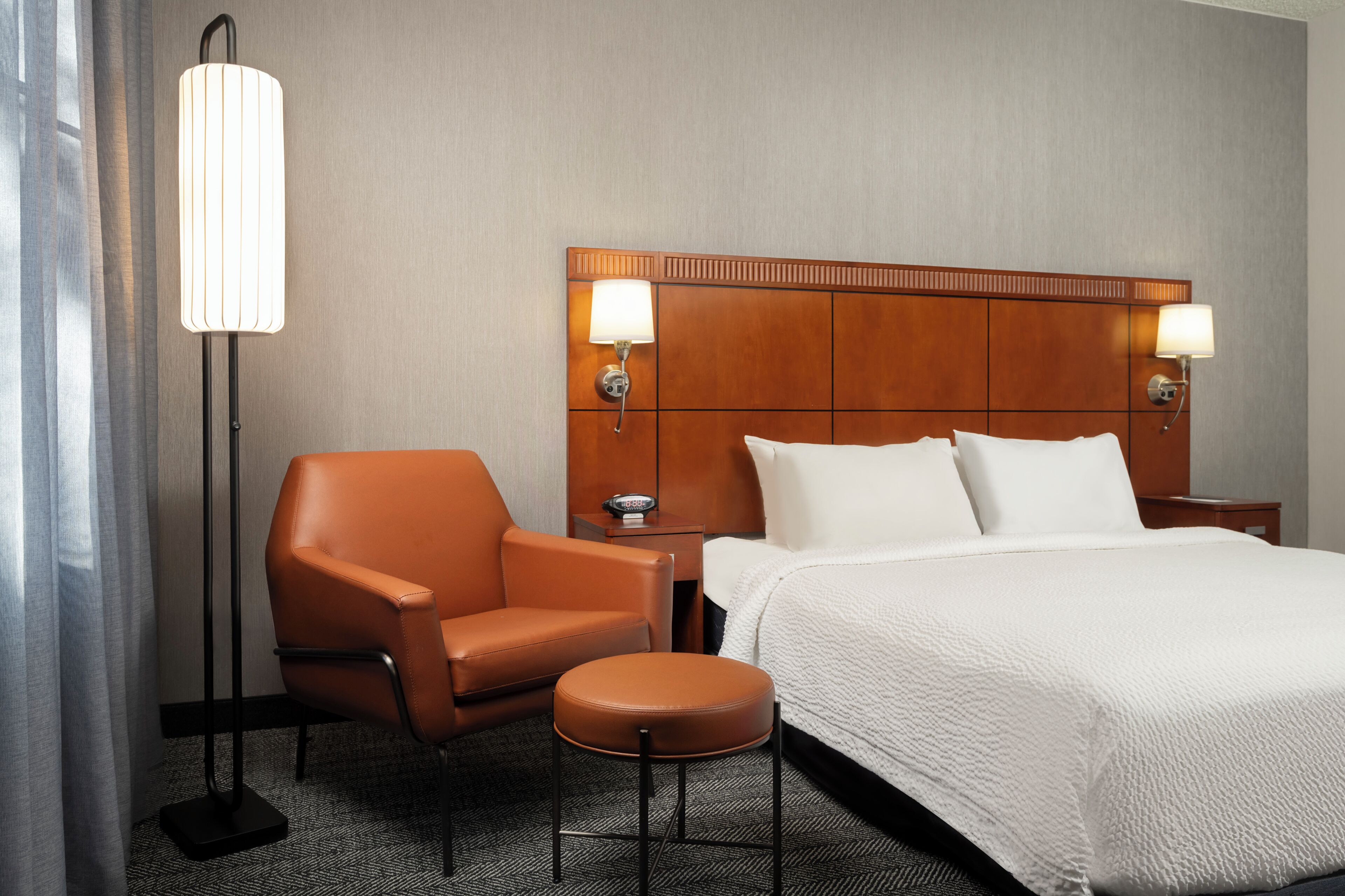 Una habitación de hotel con una cama, dos lámparas de noche, una silla, una mesita y una lámpara de pie.
