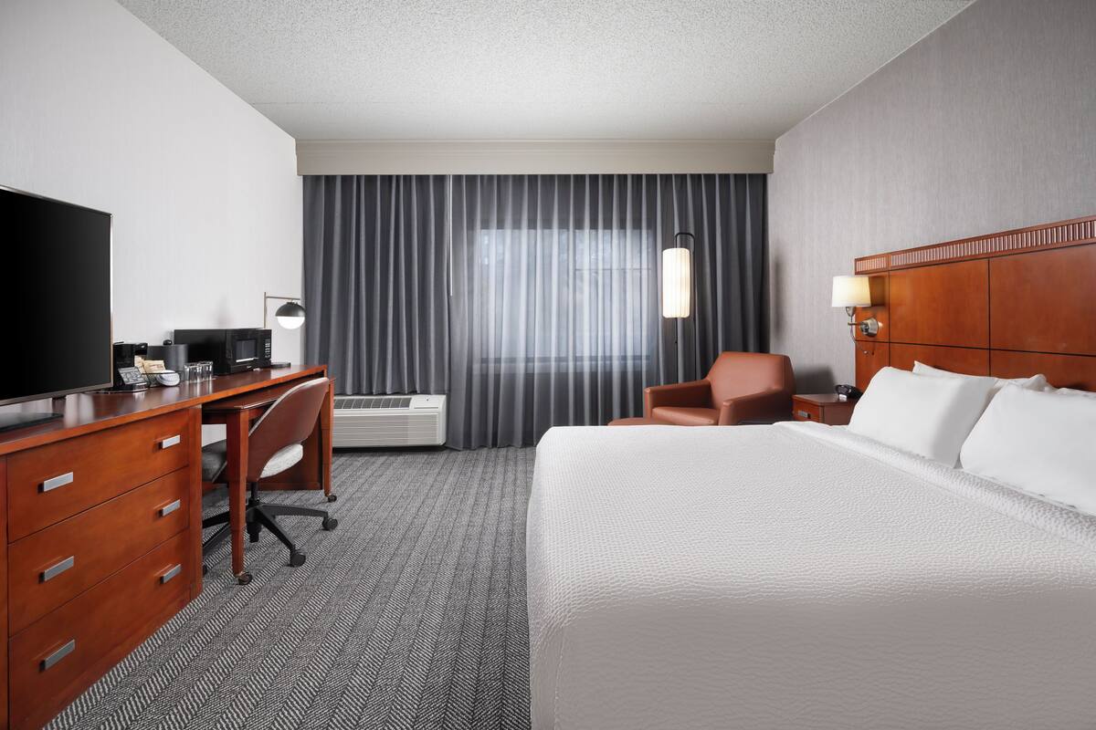 A hotel room with a bed, a desk with a computer, a chair, a television, and a window with curtains.