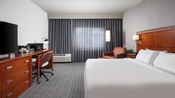 Une chambre d’hôtel comprenant un lit, un bureau avec un ordinateur, une chaise, une télévision et une fenêtre avec des rideaux.
