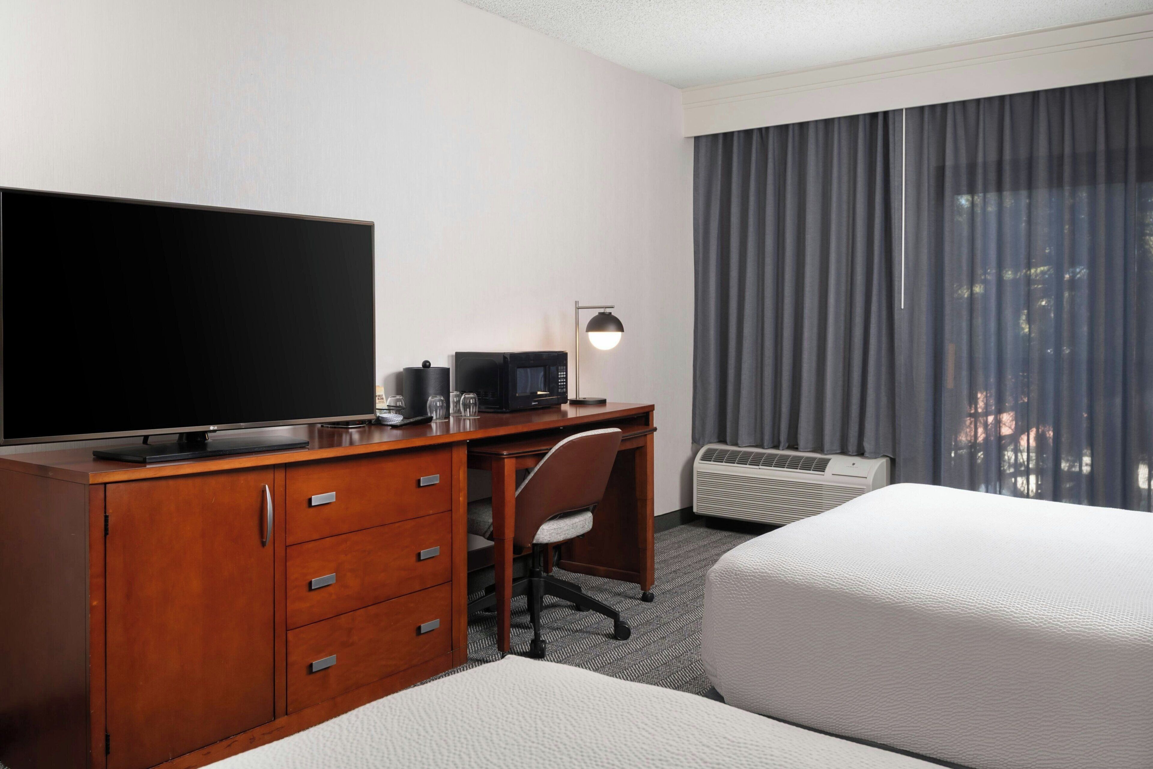 Une chambre d’hôtel équipée d’un téléviseur à écran plat, d’un bureau en bois avec des tiroirs, d’une chaise, d’un lit et d’une fenêtre avec des rideaux.