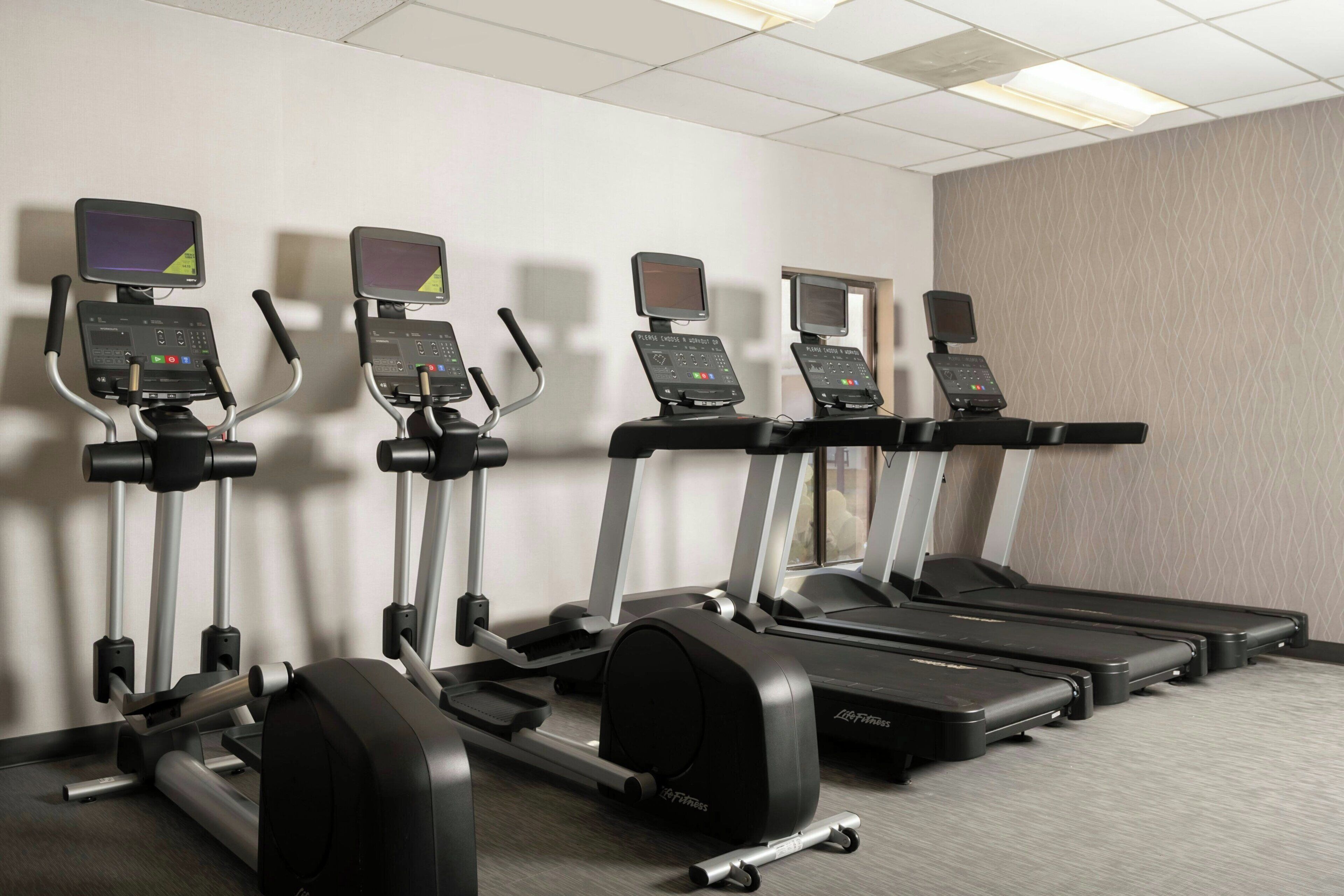 A row of treadmills in a hotel fitness room with a patterned wall and a ceiling with recessed lighting.