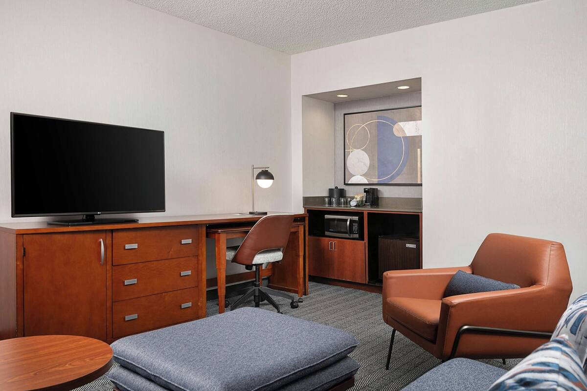 A hotel room with a flat-screen TV, wooden desk, kitchenette, and seating area.