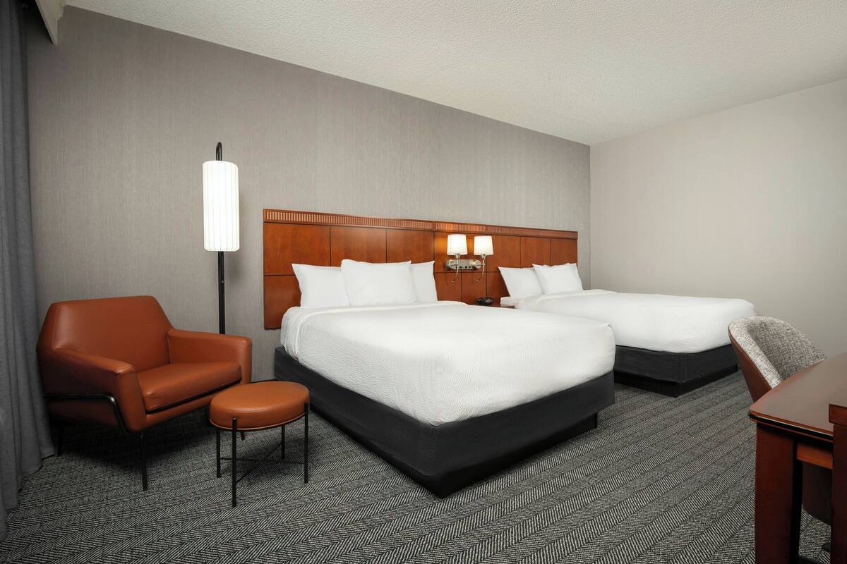 A hotel room with two beds, a chair, a desk, and a lamp.