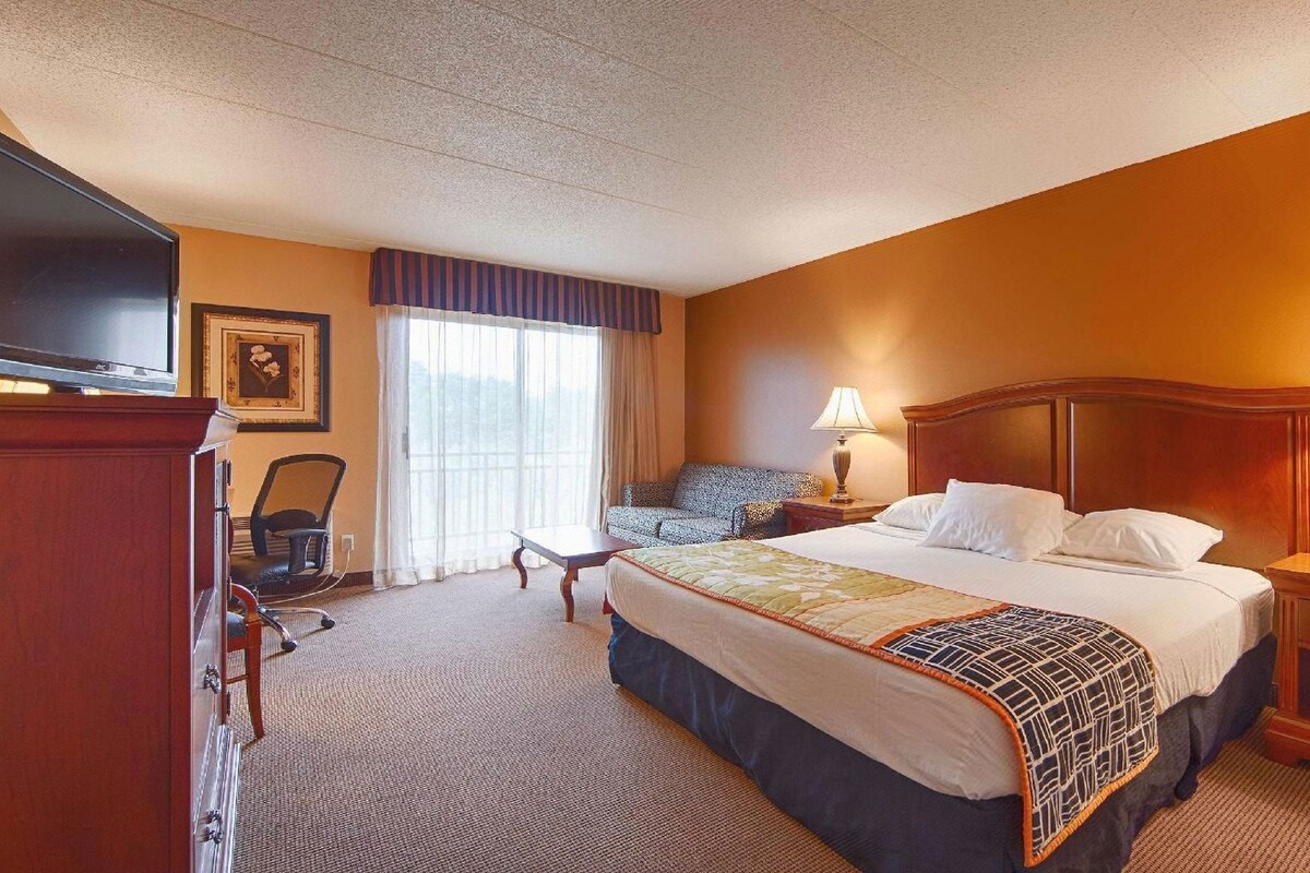 A hotel room with a bed, two bedside tables with lamps, a TV, a chair, a desk, and a window with curtains.