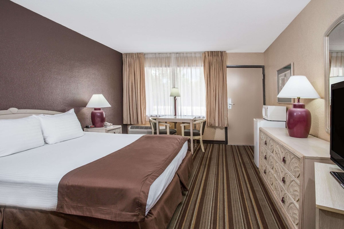 Une chambre d’hôtel avec un grand lit, un bureau avec une chaise, une télévision, une commode et une fenêtre avec des rideaux.
