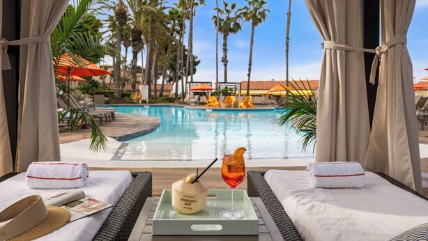 A poolside view with a lounge chair, a towel, a tray with a drink and a coconut, and a pool with palm trees in the background. - San Diego Mission Bay Resort (San Diego)