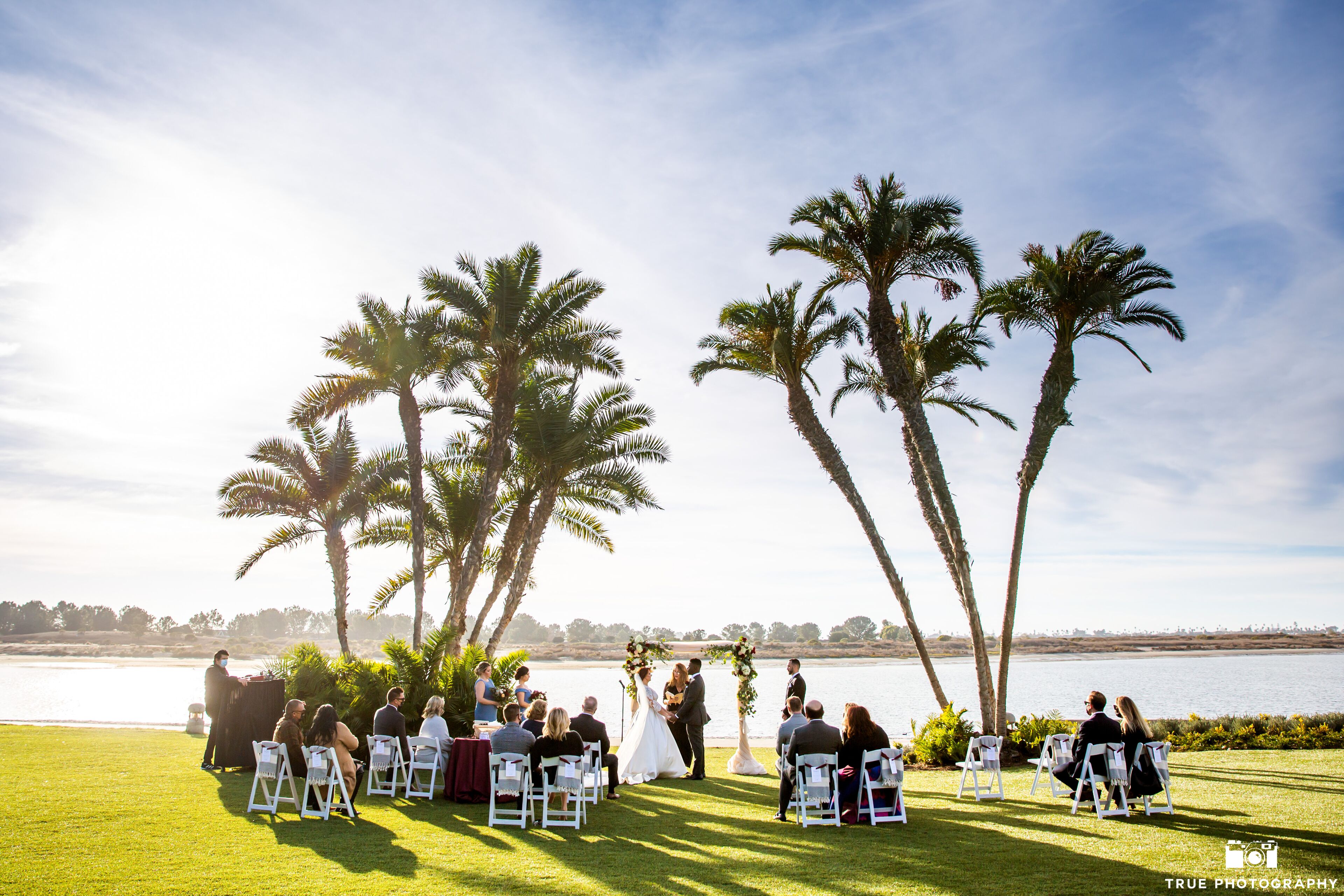 A wedding ceremony outdoors with guests seated on white chairs, palm trees, and a body of water in the background.