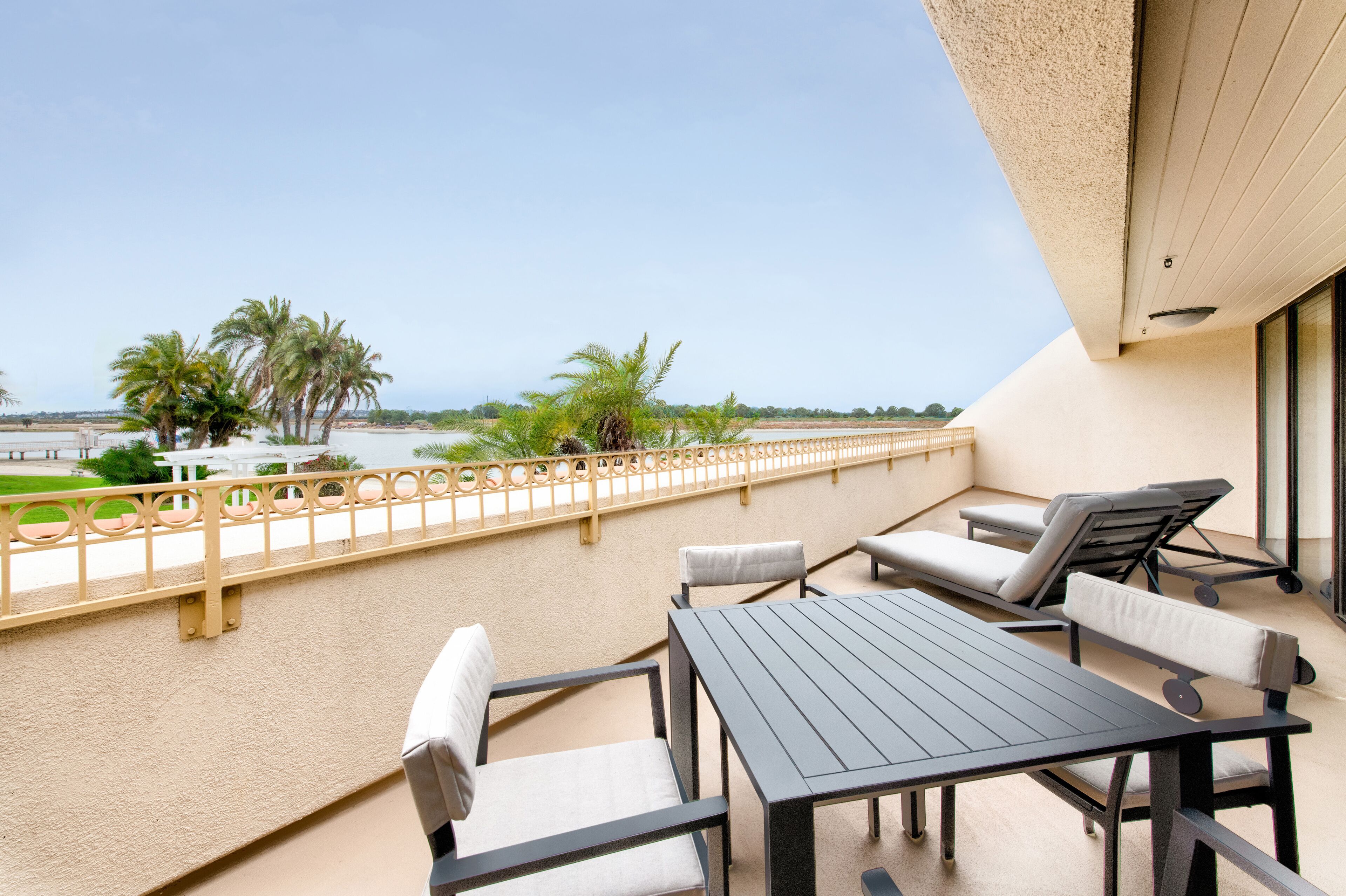 A balcony with a table and chairs, a view of the beach, and palm trees.