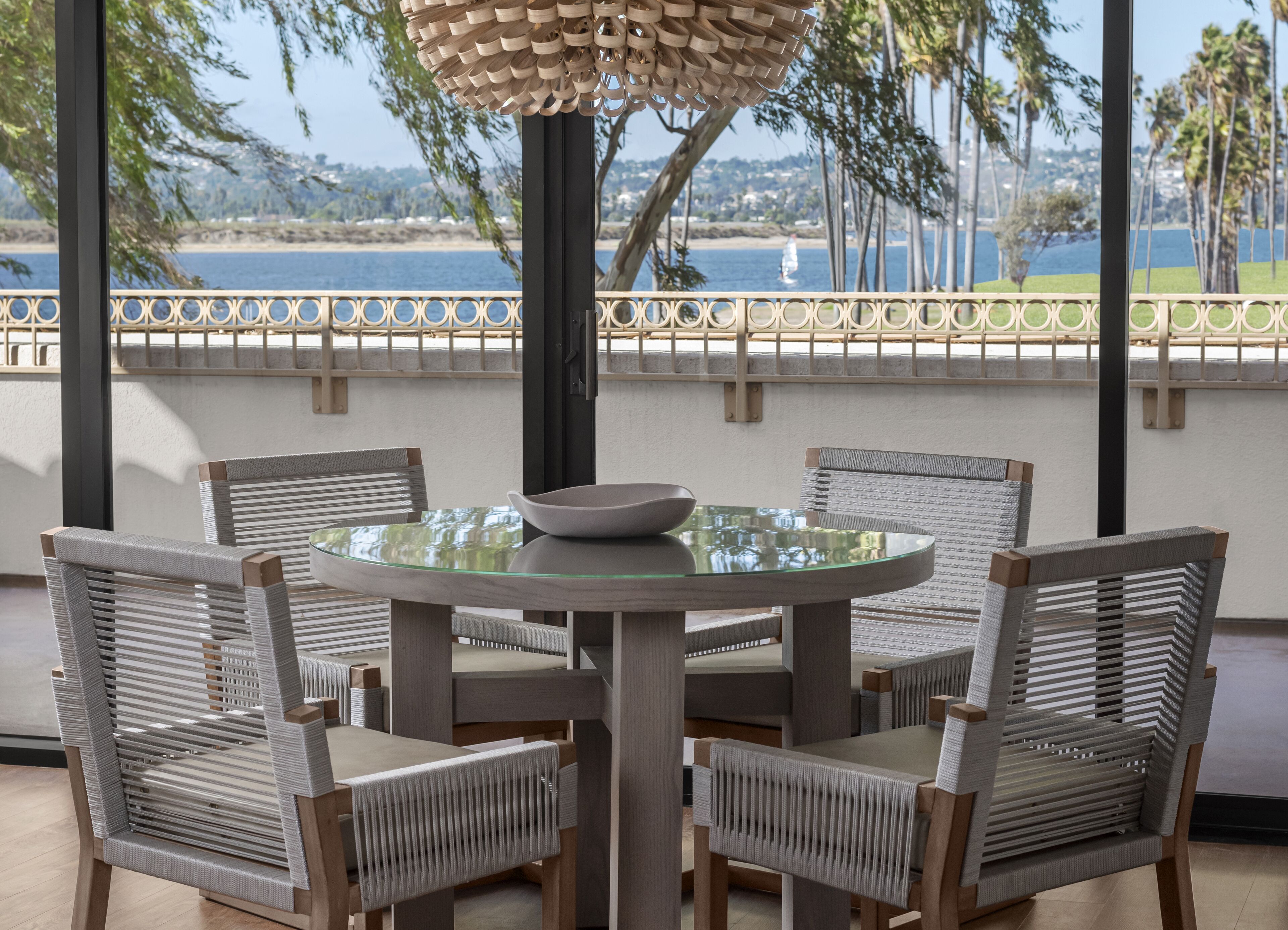Mesa de vidro redonda com quatro cadeiras de vime, um guarda-sol de madeira decorativo e vista para um lago com um veleiro.