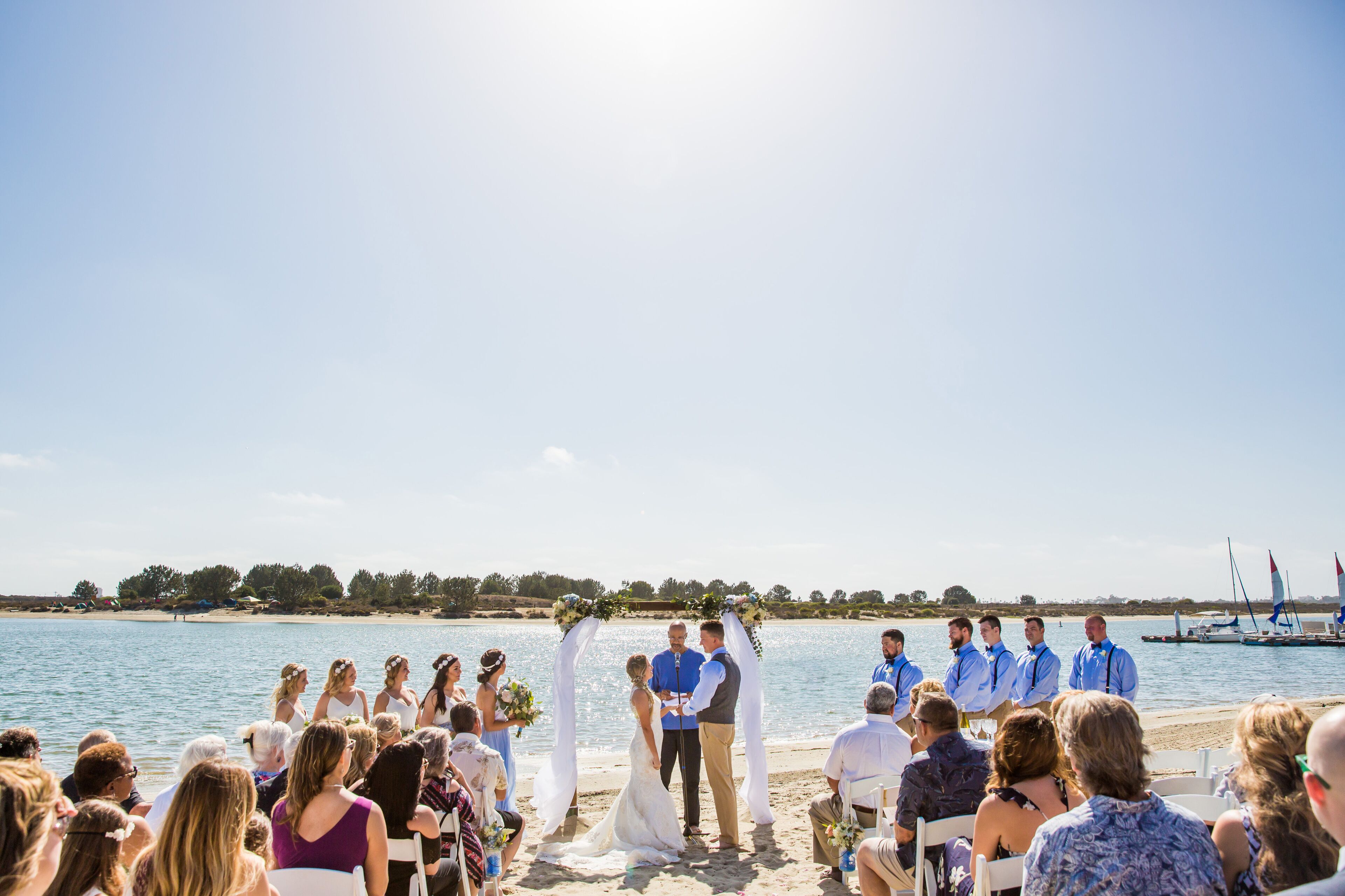 A beach wedding ceremony with guests seated facing the altar, a clear sky, and a calm sea in the background.