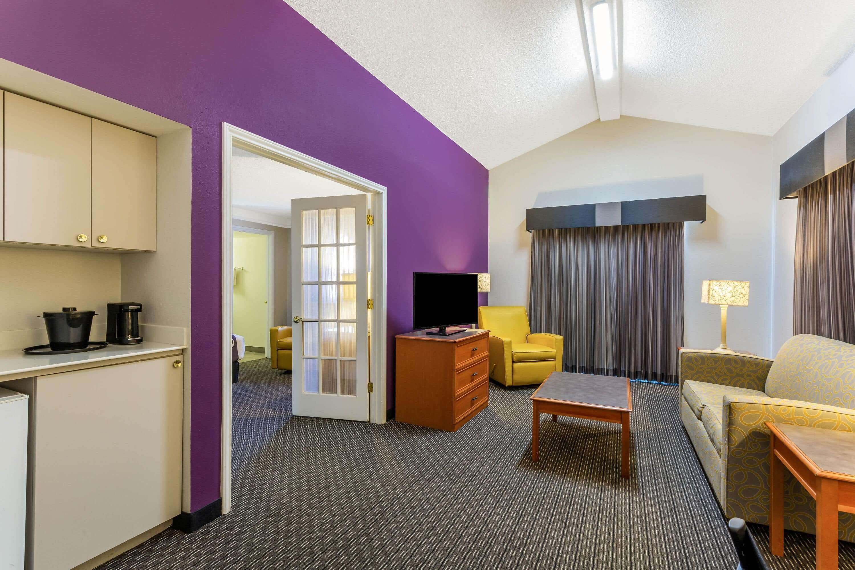 A hotel room with a purple accent wall, a kitchenette with a fridge, a TV, a yellow armchair, a sofa, and a lamp.