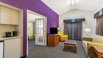 A hotel room with a purple accent wall, a kitchenette with a fridge, a TV, a yellow armchair, a sofa, and a lamp.