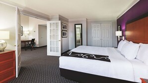 Habitación de hotel con una cama grande, un escritorio y una silla. Se observa una puerta que da a otra habitación y una lámpara fijada a la pared.