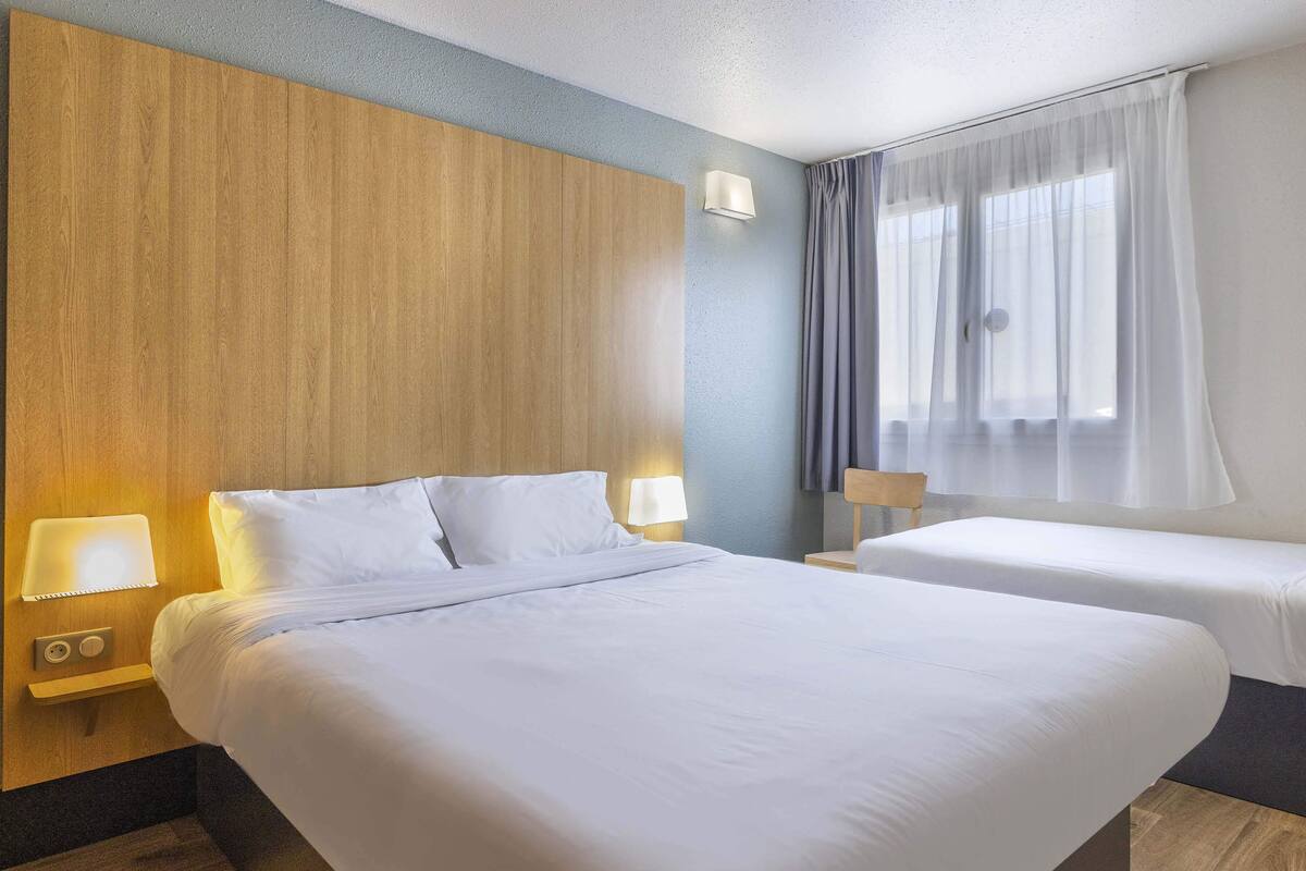Een hotelkamer met een groot bed, een kleiner bed, een nachtkastje met een lamp en een raam met gordijnen.