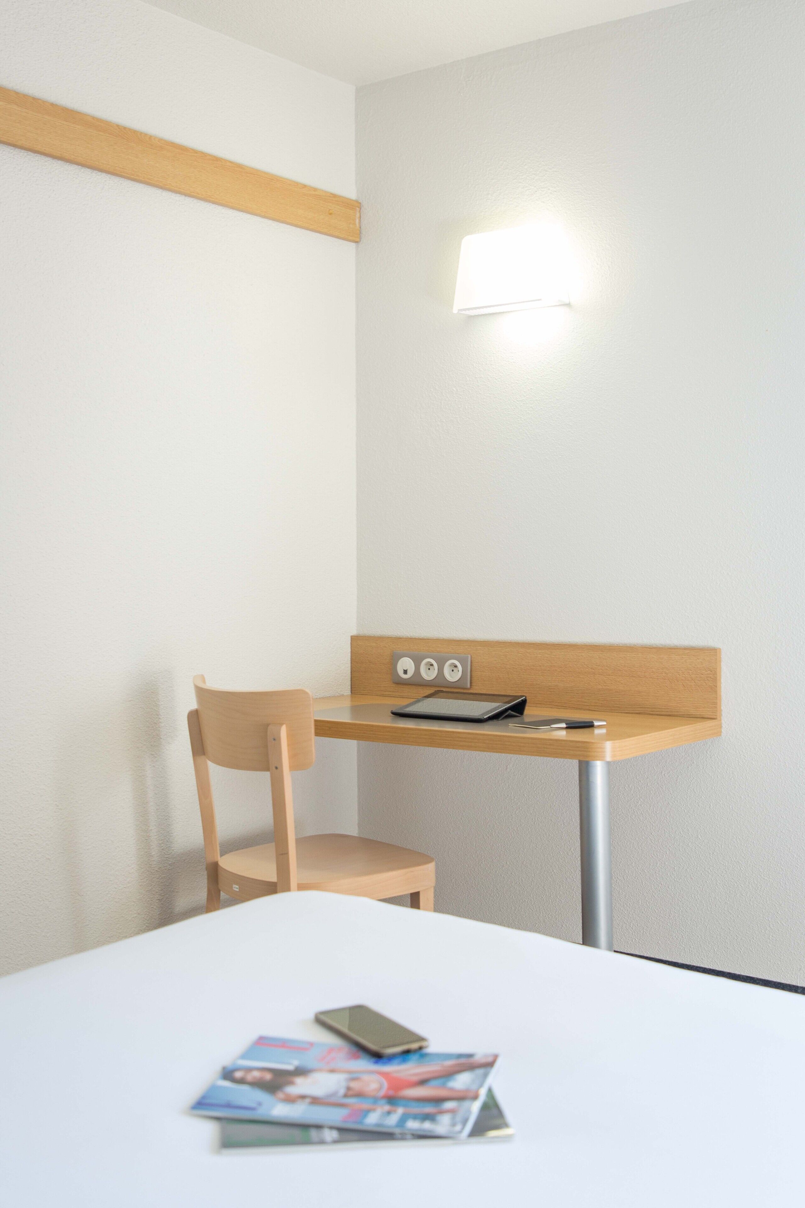 Una habitación de hotel pequeña y ordenada, con una cama individual, un escritorio de madera, una silla y una mesita de noche.