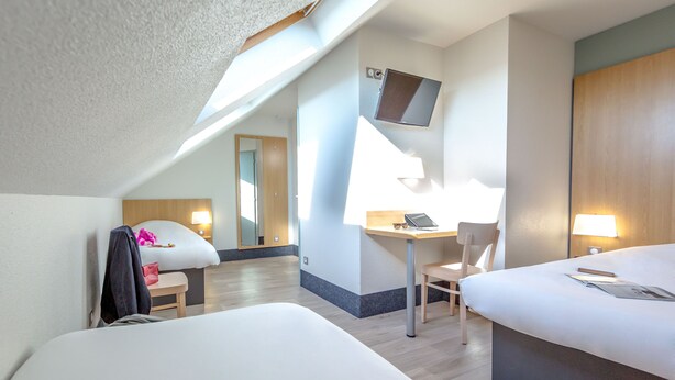 A hotel room with a bed, a desk with a chair, a TV mounted on the wall, and a window providing natural light. - B&B HOTEL Perpignan Nord (Perpignan)