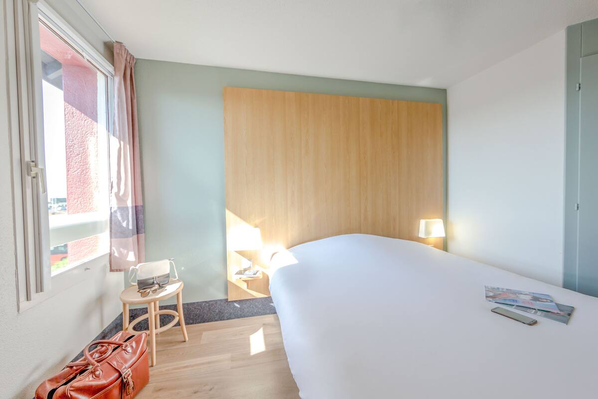 Een hotelkamer met een groot bed, een klein tafeltje met een stoel, een lamp en een handtas op de vloer.