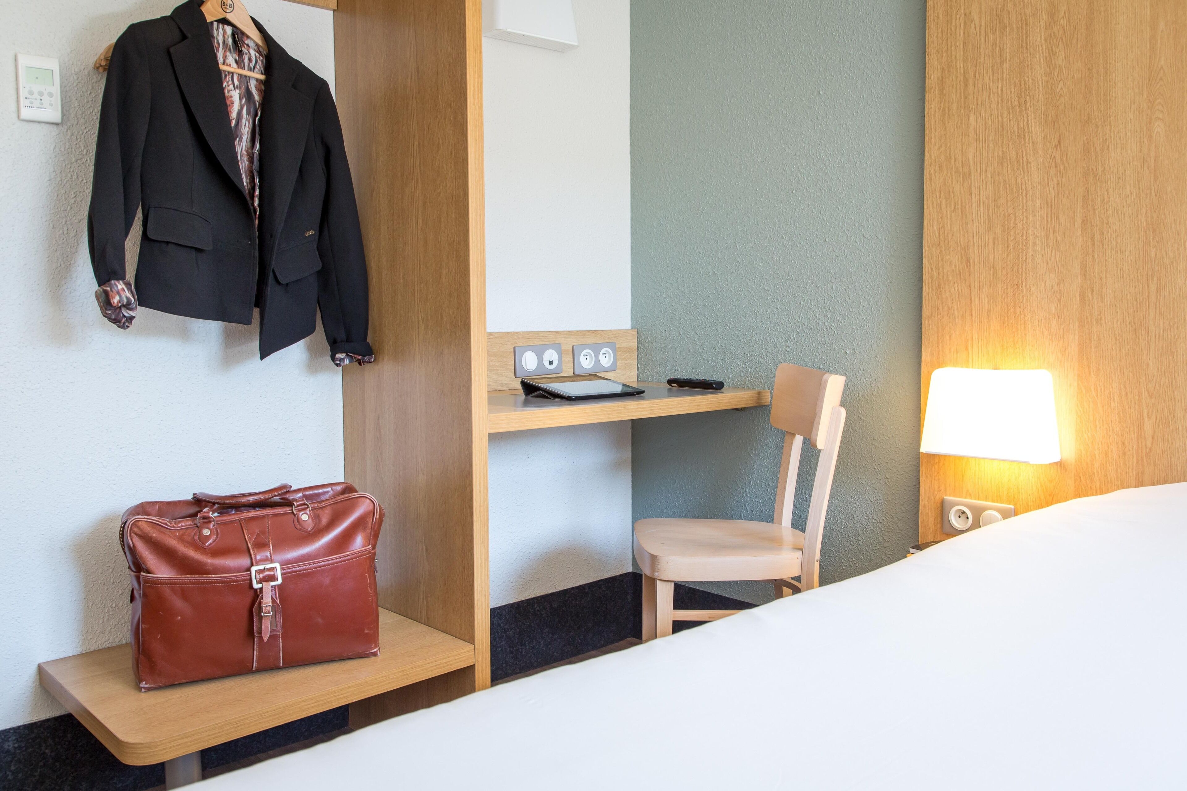 Une chambre d’hôtel avec un lit, un bureau et une chaise, un manteau accroché et un sac à main posé sur une étagère.