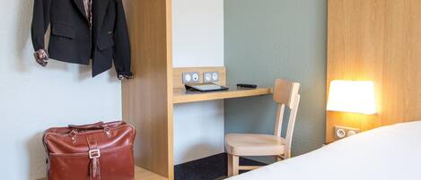 Habitación de hotel con una cama, un escritorio con silla, un abrigo colgado y un bolso en una estantería.
