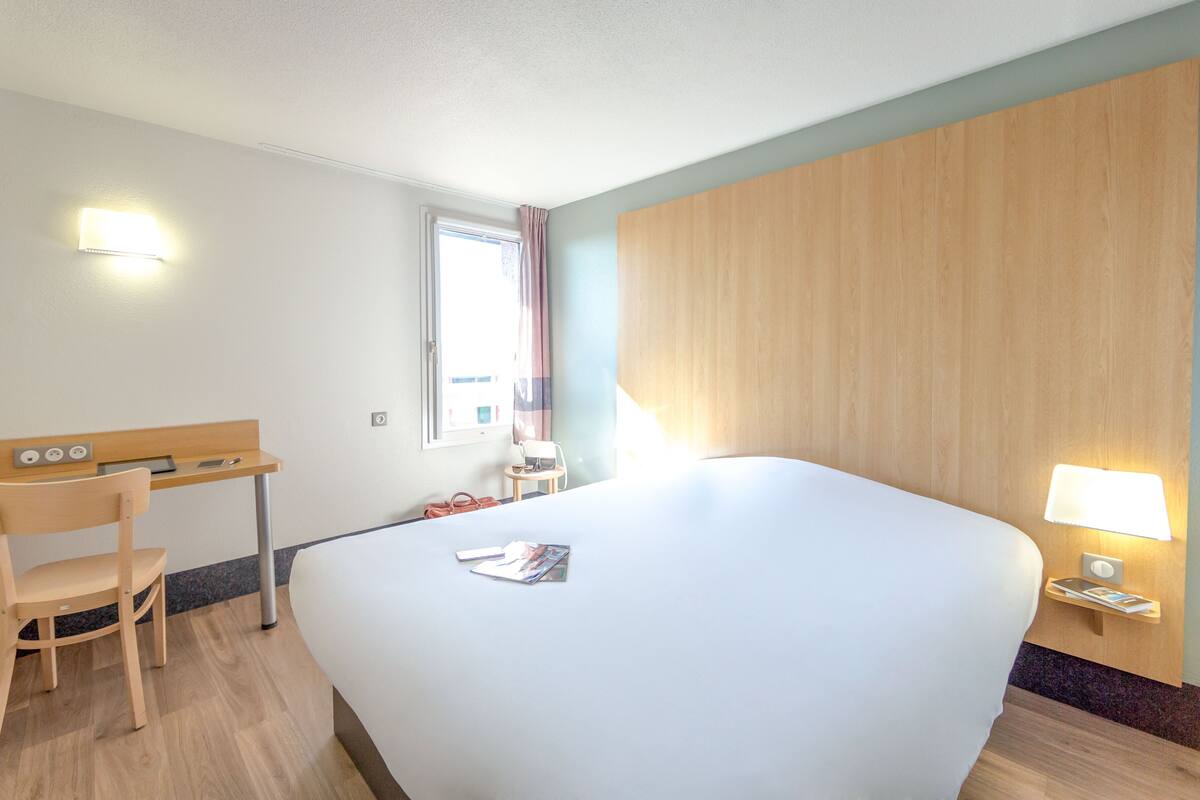 Een hotelkamer met een groot bed, een bureau met een stoel, een kleine tafel met een lamp en een raam met gordijnen.