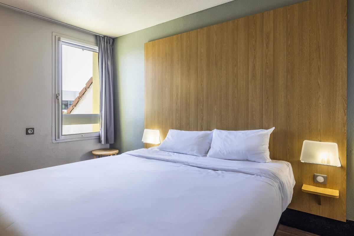 Een net opgemaakt bed met witte lakens, een houten hoofdbord, twee bedlampjes en een raam met gordijnen.