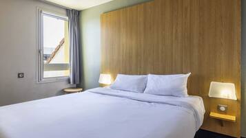 A neatly made bed with white linens, a wooden headboard, two bedside lamps, and a window with curtains.