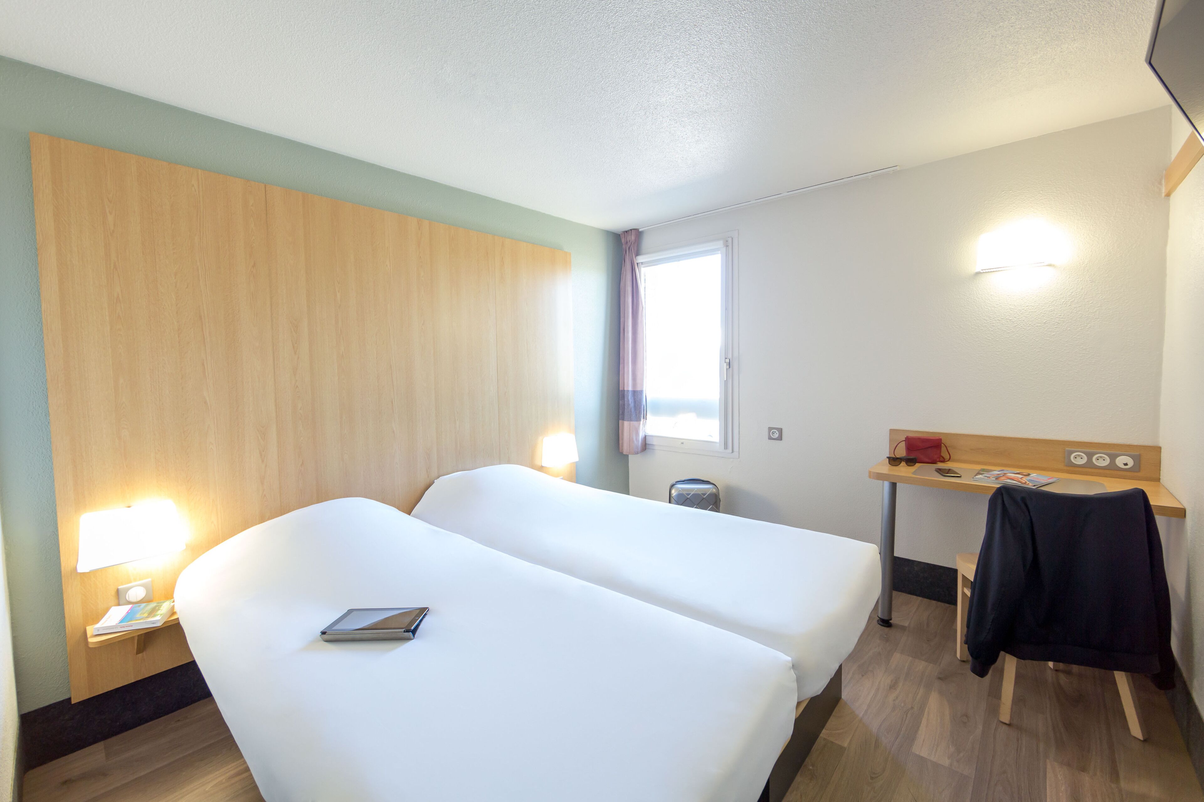 Ein Hotelzimmer mit zwei Betten, einem Schreibtisch, einer Lampe und einem Fenster.