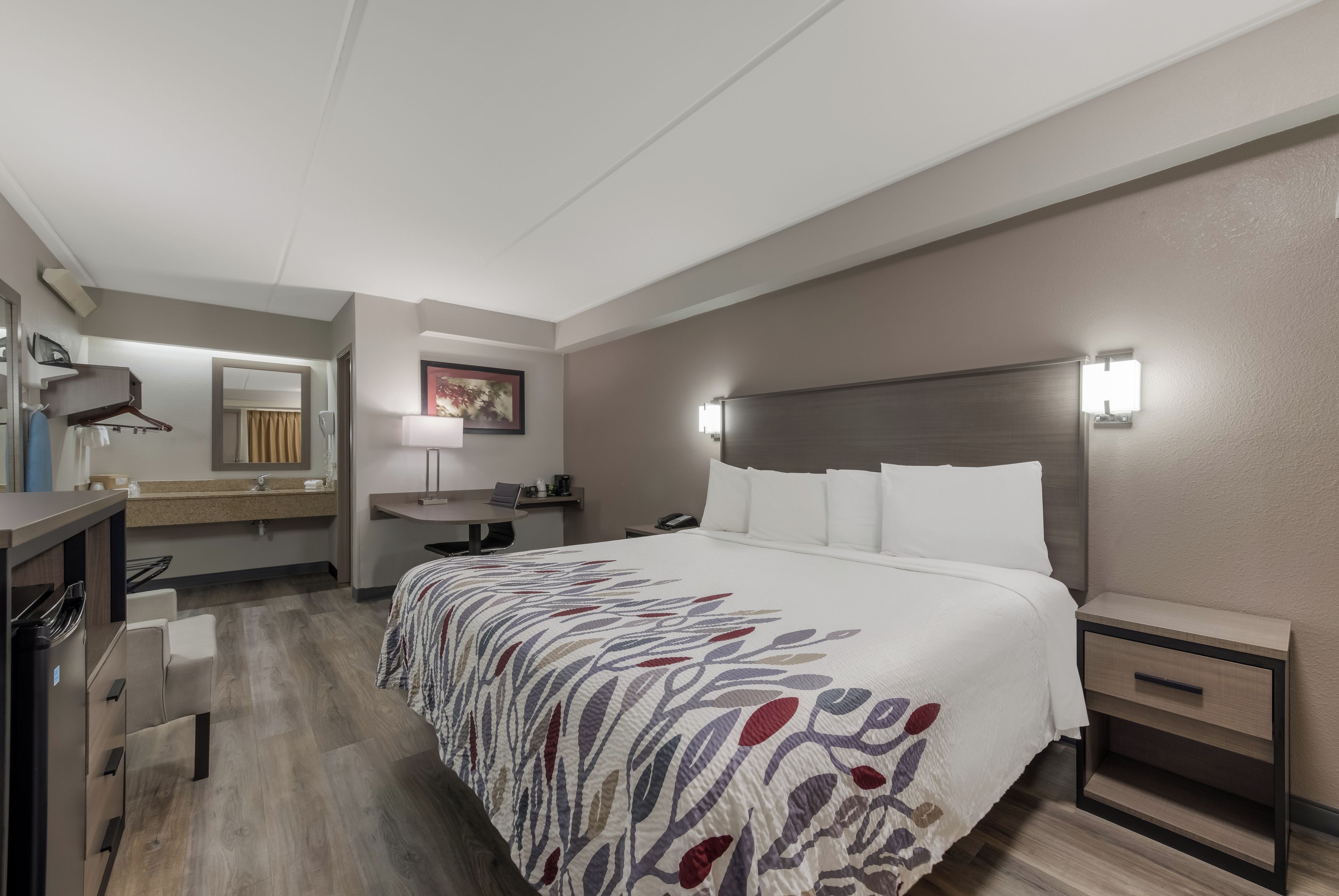 A hotel room with a bed, bedside table, nightstand, desk, chair, and a bathroom with a sink and mirror.