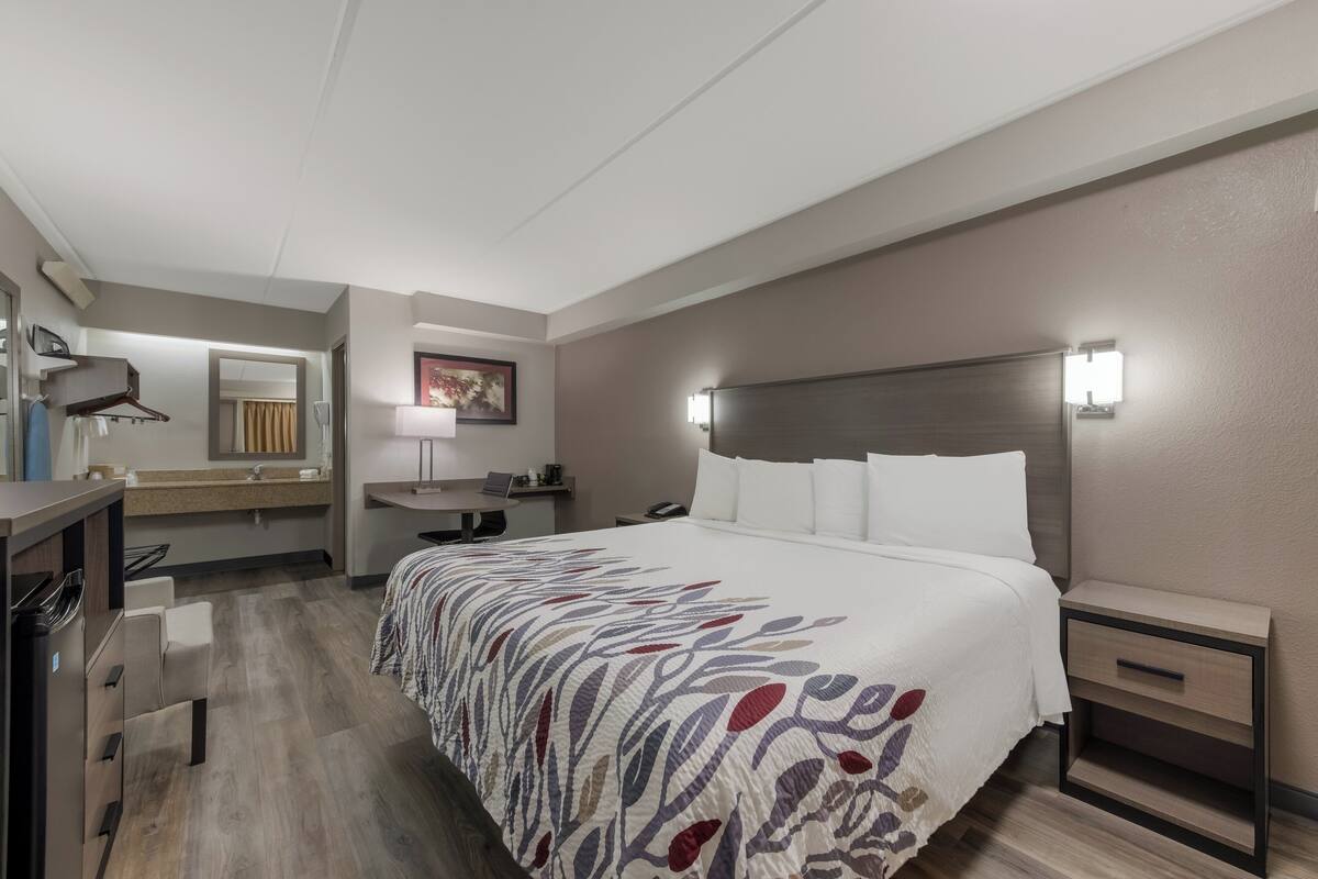 A hotel room with a bed, bedside table, nightstand, desk, chair, and a bathroom with a sink and mirror.