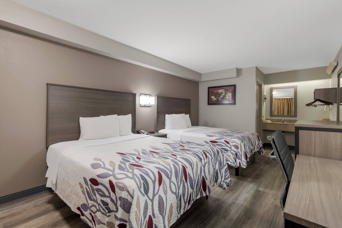 A hotel room with two beds, a desk, a chair, a mirror, and a bathroom visible through an open door.