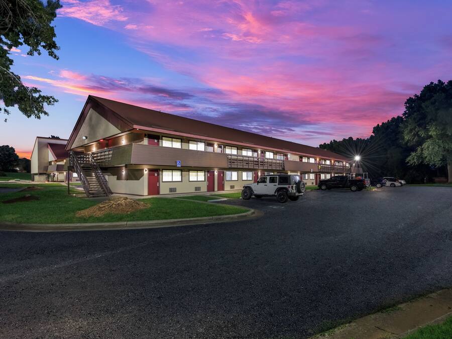 A motel with a red roof, multiple windows, and a parking area with vehicles.