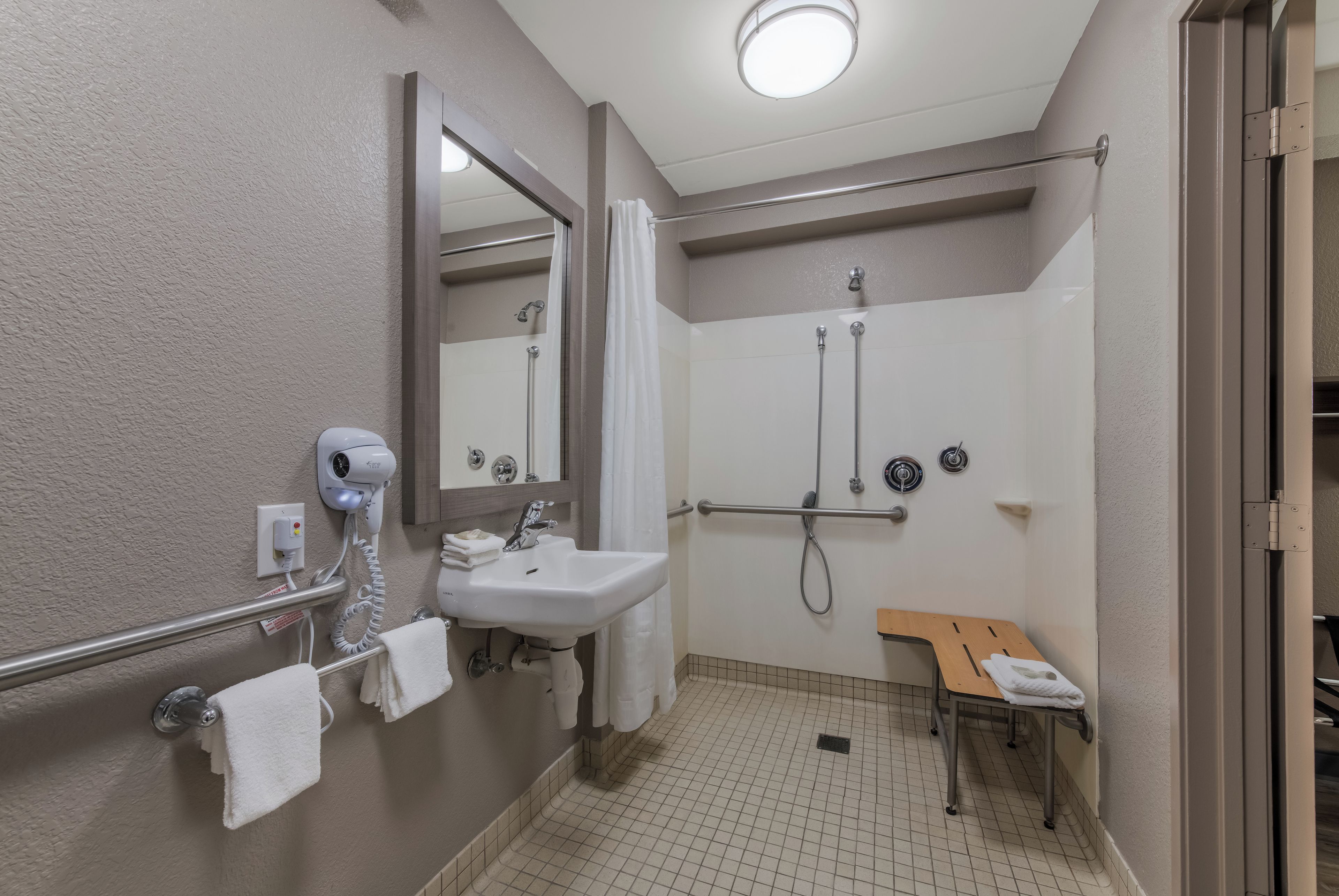 Ein Badezimmer mit einem weißen Waschbecken, einem Spiegel, einer Handbrause, einem Handtuchhalter und einer Sitzbank.