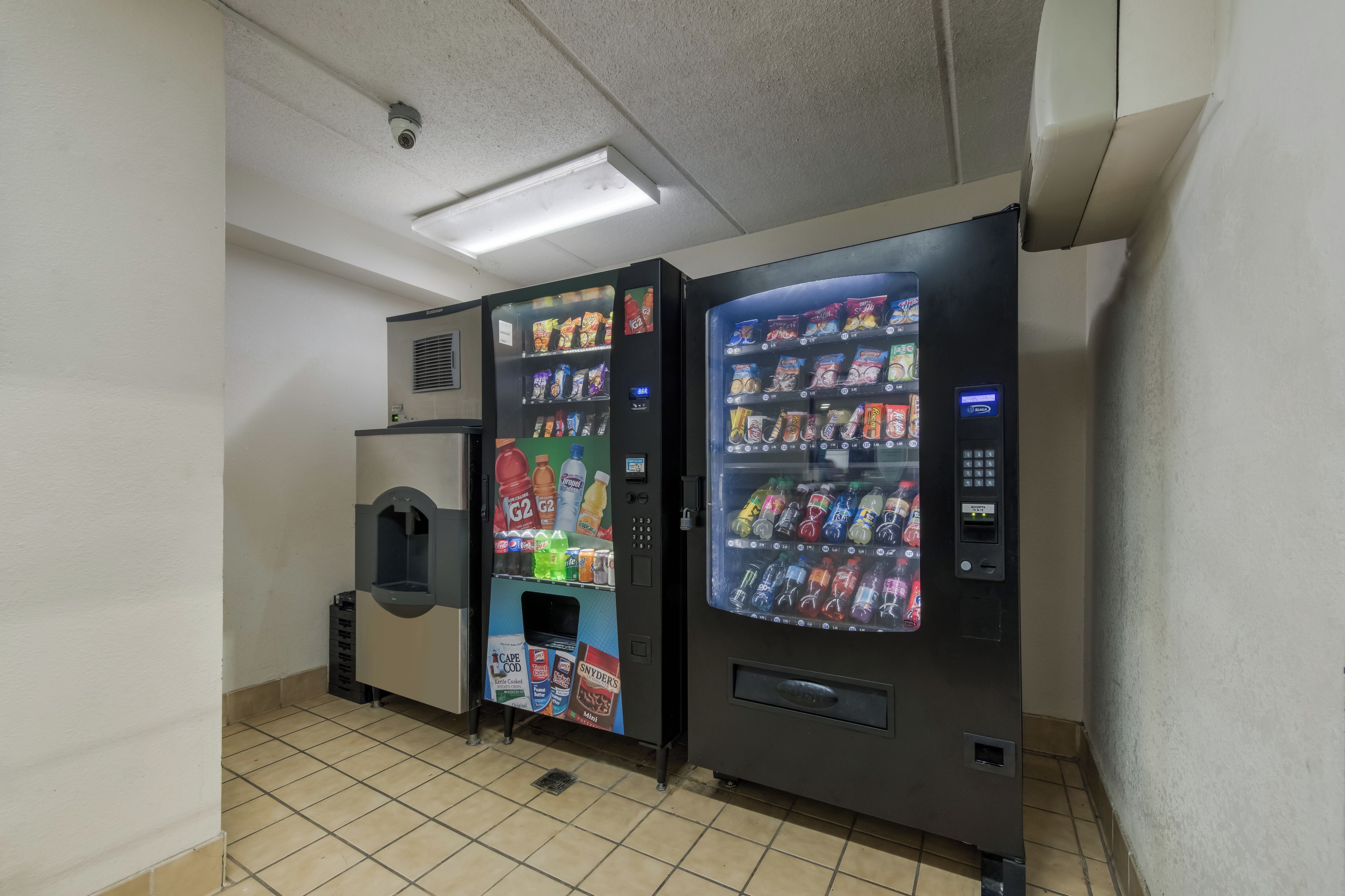 Two vending machines with a variety of snacks and drinks, a security camera, and a tiled floor.