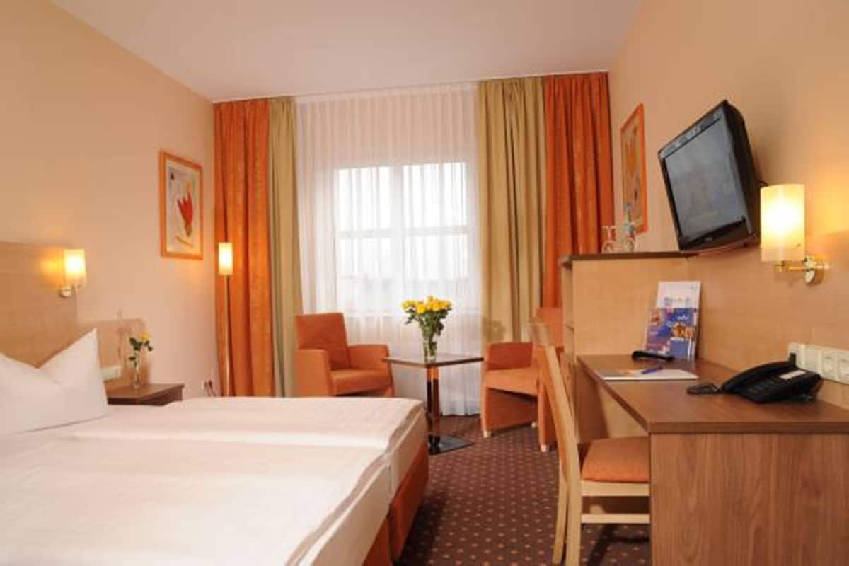 A hotel room with a bed, a desk, a chair, a television, and a small table with a vase of flowers.