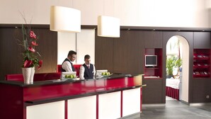A reception desk with two staff members, a large vase with flowers, and a television on a shelf. - PLAZA INN Berlin Charlottenburg (Berlin)