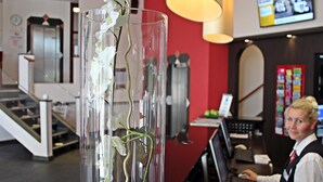 A reception area with a glass vase of white orchids, a desk with a computer, and a red wall with a TV screen. - PLAZA INN Berlin Charlottenburg (Berlin)