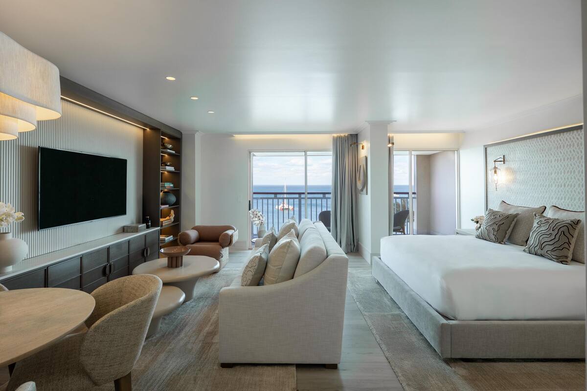A modern hotel room with a large bed, a sofa, a TV, a dining area, and a view of the sea.