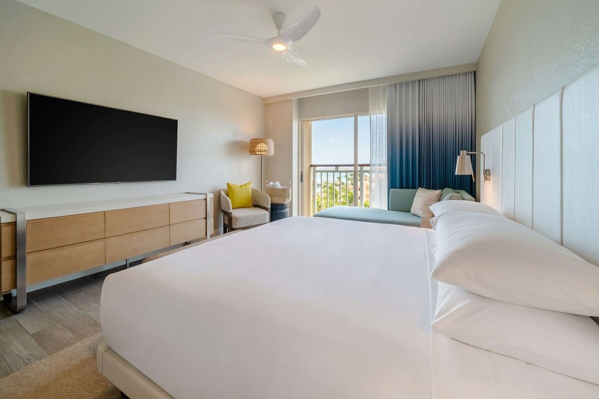 Une chambre d’hôtel dotée d’un grand lit, d’un téléviseur à écran plat, d’un bureau avec une chaise, d’un balcon avec vue et d’un ventilateur de plafond.
