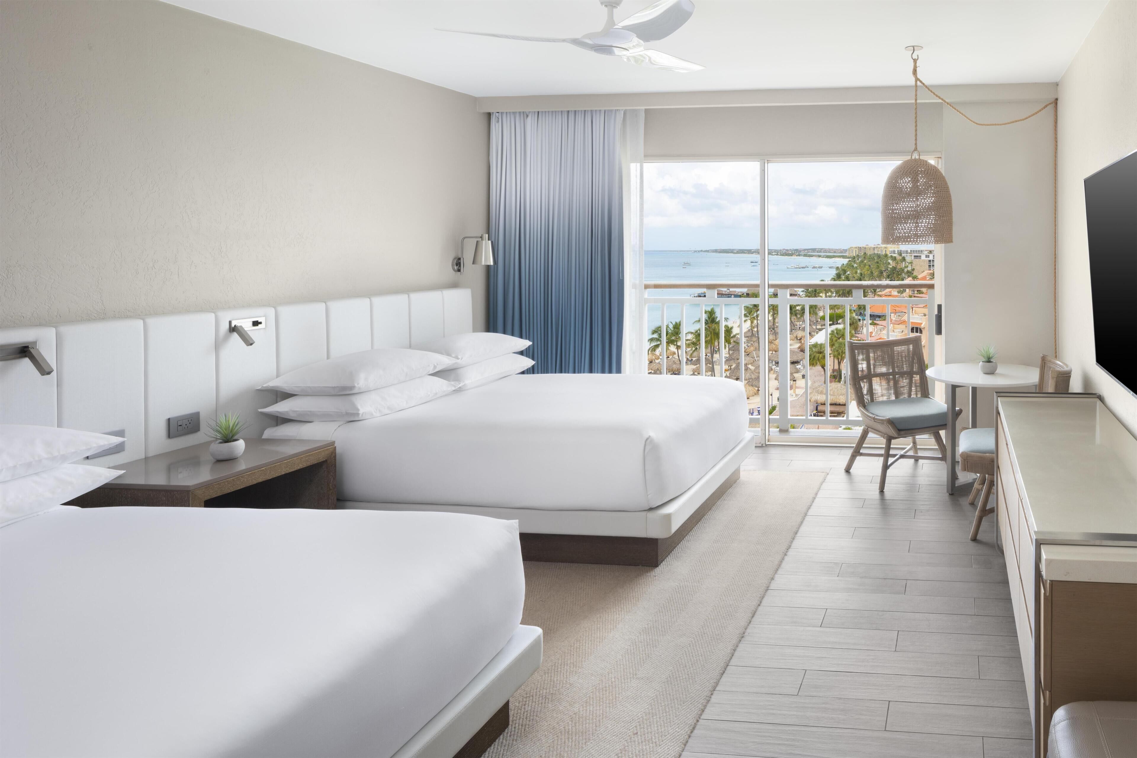A hotel room with two beds, a desk, a chair, a TV, a balcony with a view of the sea, and a ceiling fan.