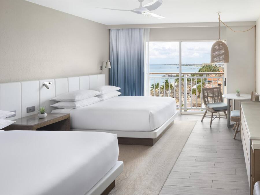 A hotel room with two beds, a desk, a chair, a TV, a balcony with a view of the sea, and a ceiling fan.