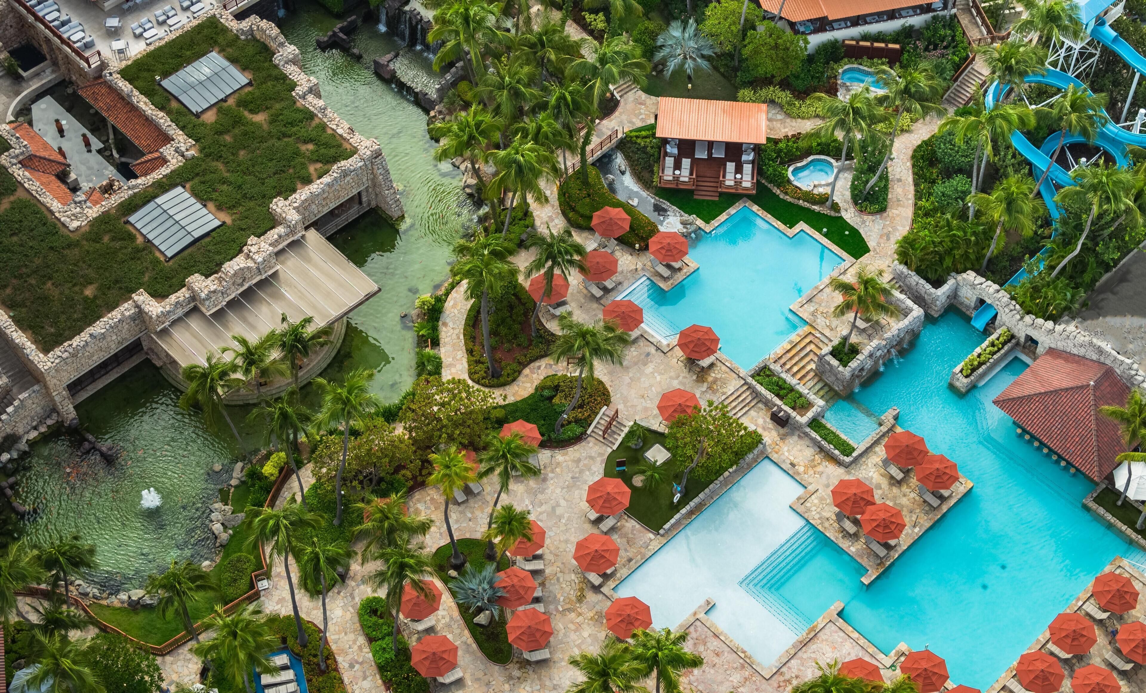Vista aérea de un complejo turístico con varias piscinas, palmeras y senderos de piedra.