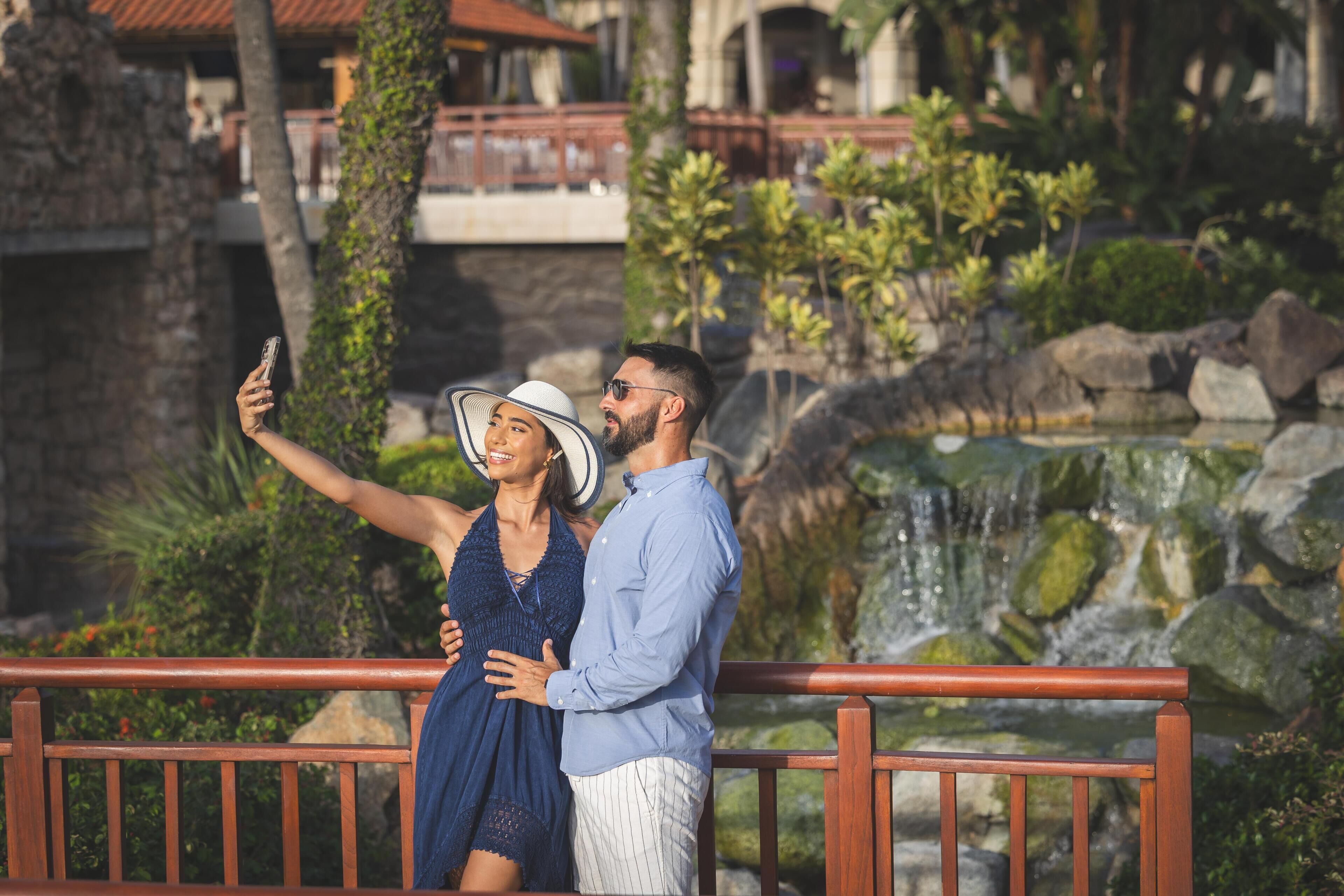 A couple taking a selfie on a wooden bridge with a waterfall and greenery in the background.
