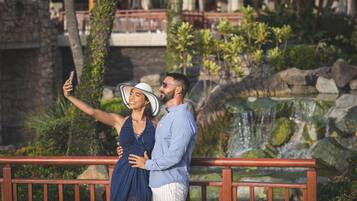 A couple taking a selfie on a wooden bridge with a waterfall and greenery in the background.