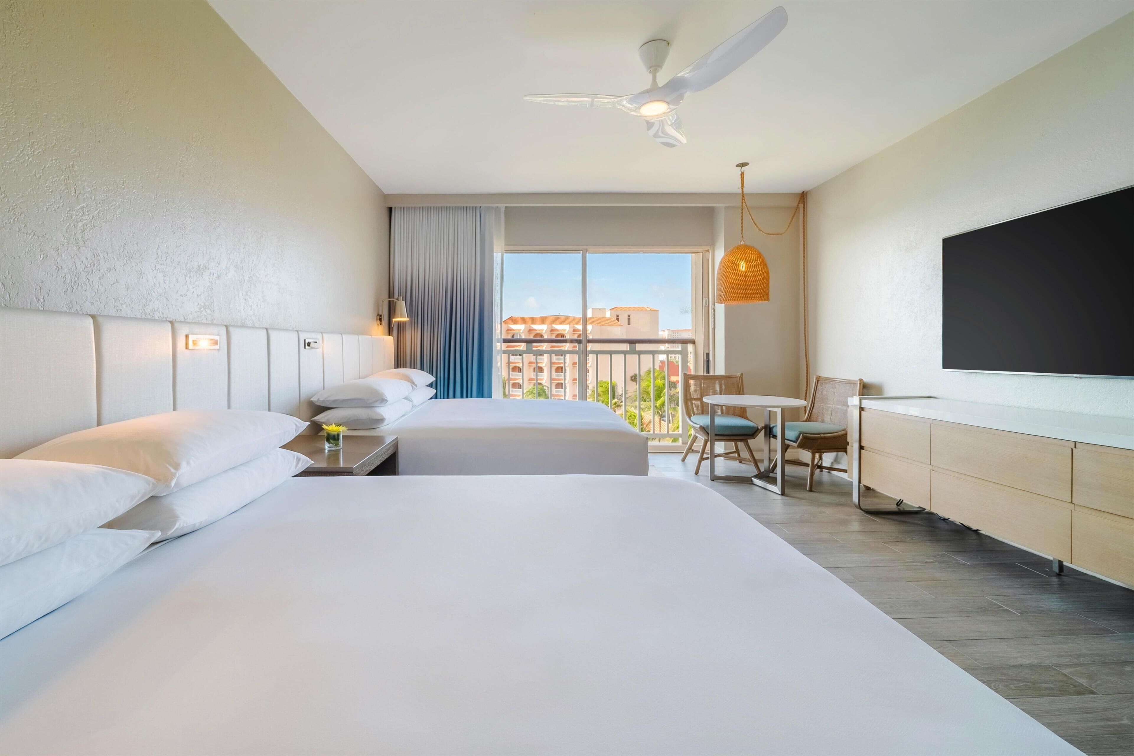 Quarto de hotel com duas camas, uma mesa, uma cadeira, televisão, ventoinha no teto e varanda com vista para edifícios.