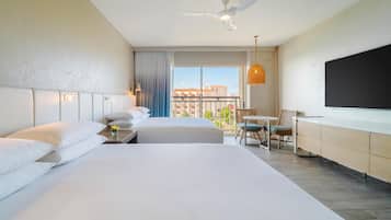 Quarto de hotel com duas camas, uma mesa, uma cadeira, televisão, ventoinha no teto e varanda com vista para edifícios.