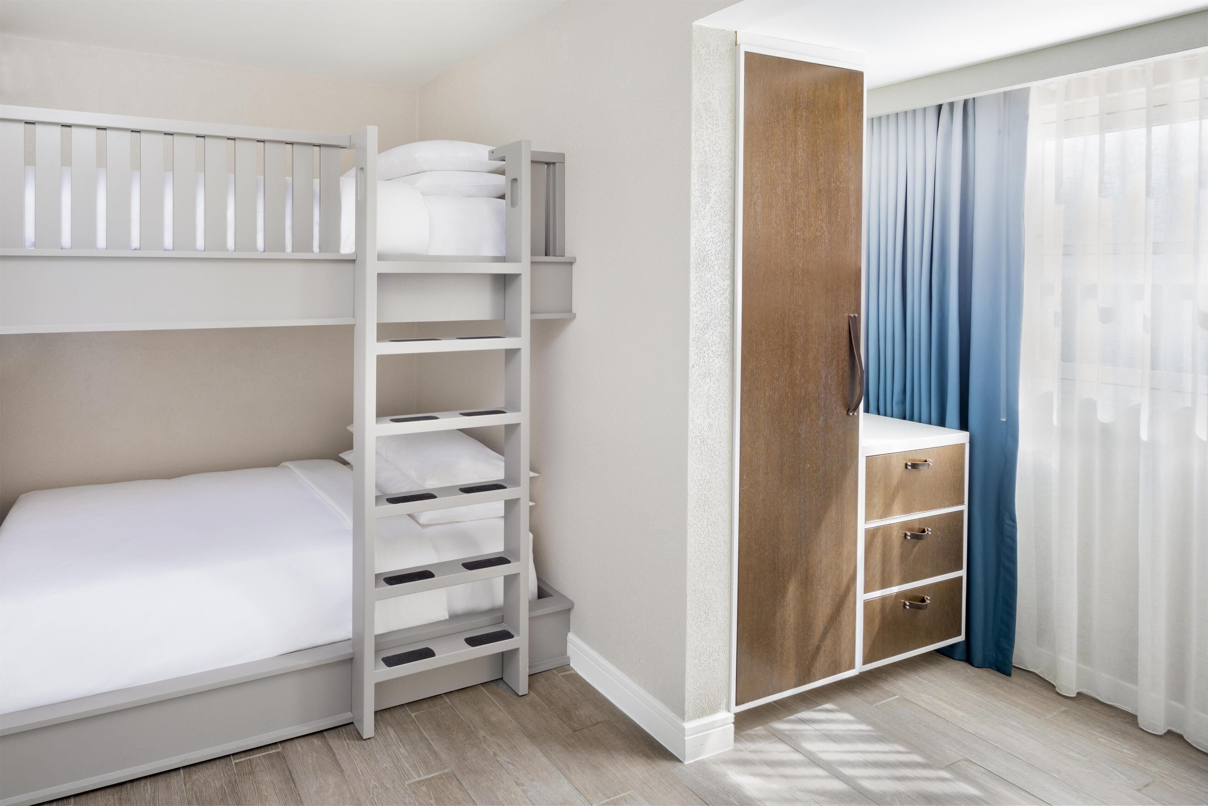 Un lit superposé avec des tiroirs de rangement, une armoire en bois et une fenêtre avec des rideaux légers.